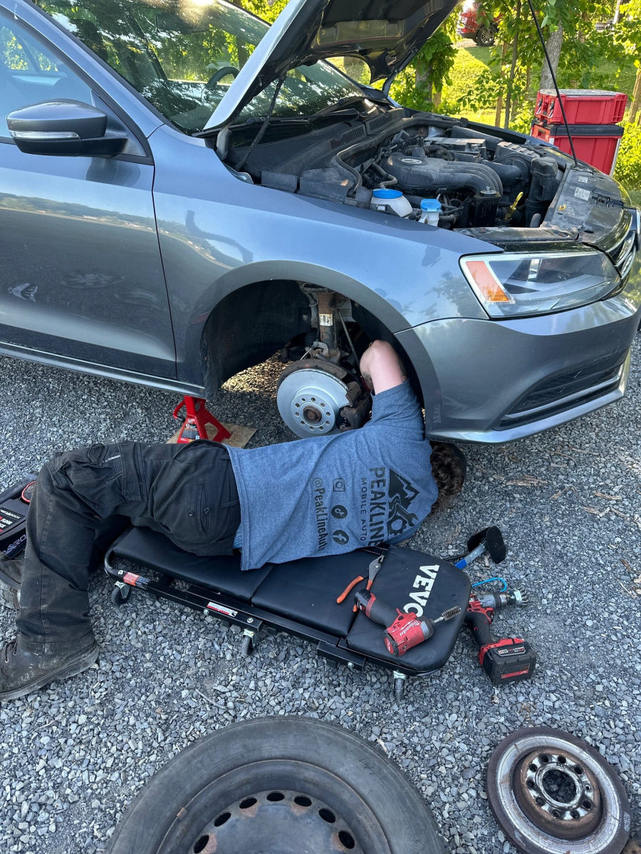 A technician on a creeper under a Volkswagen with the front wheel off, doing brakes on a driveway