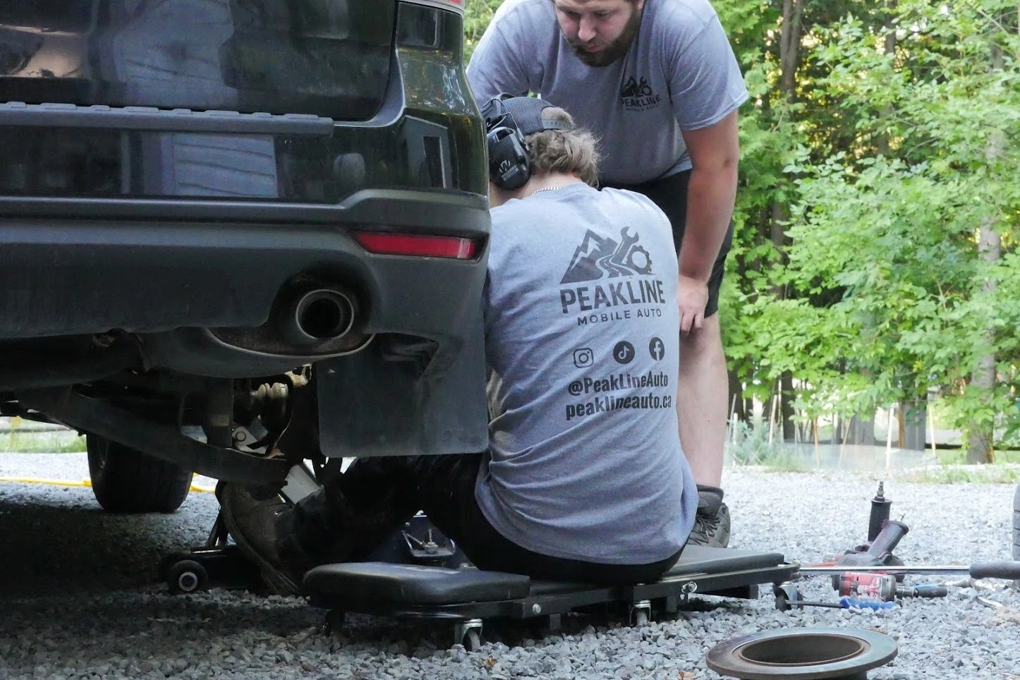 Two Peakline technicians working under the rear of an SUV on a gravel driveway