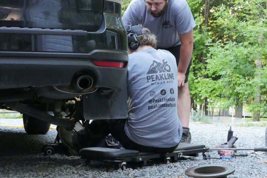 Two Peakline technicians working under the rear of an SUV on a gravel driveway
